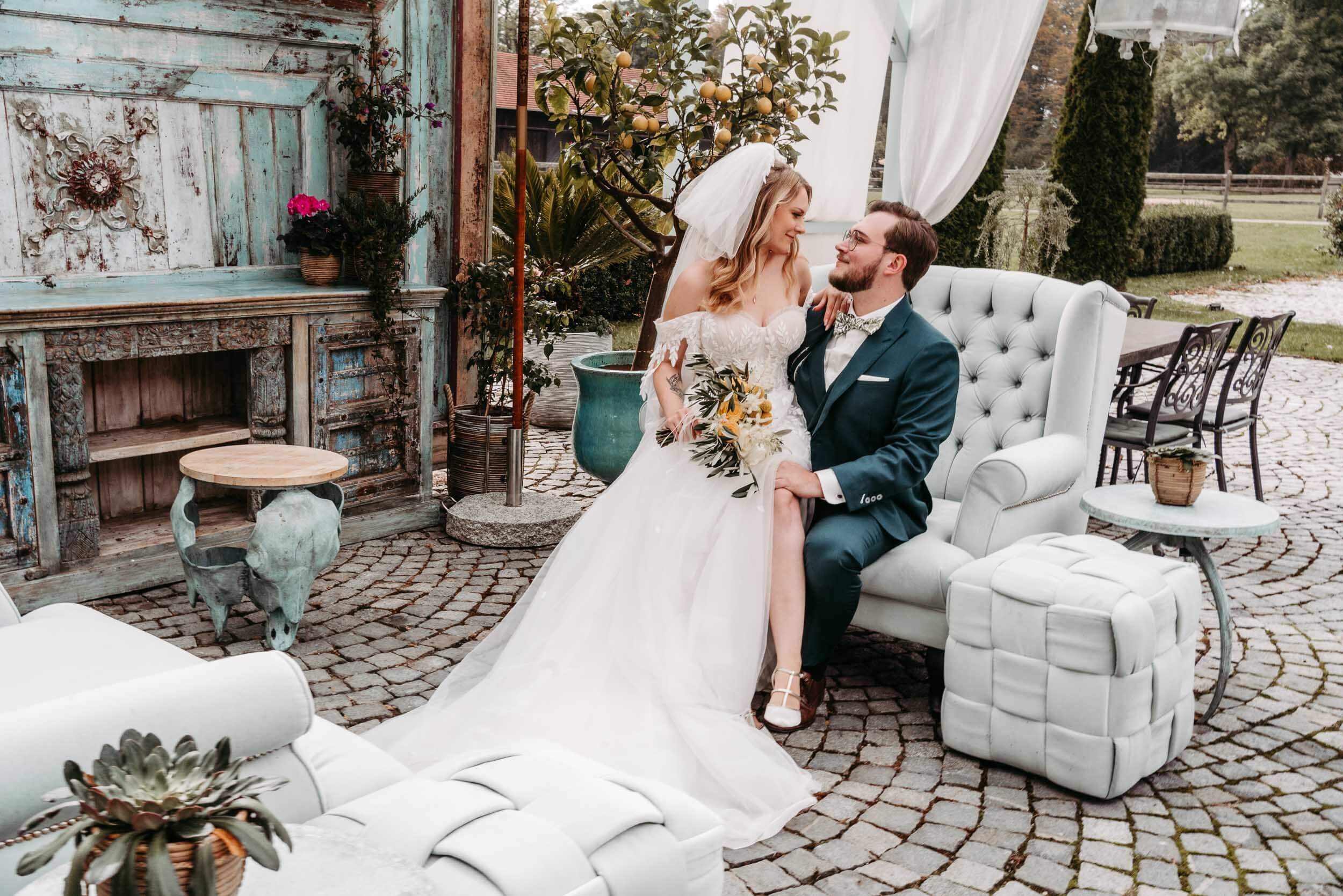 Eine Braut in einem weißen Kleid und Schleier sitzt auf dem Schoß eines Bräutigams in einem blauen Anzug. Die beiden lächeln sich an, umgeben von eleganter Außendekoration im Vintage-Stil und Grünpflanzen, die das bezaubernde Gefühl von Magical Homes vermitteln.