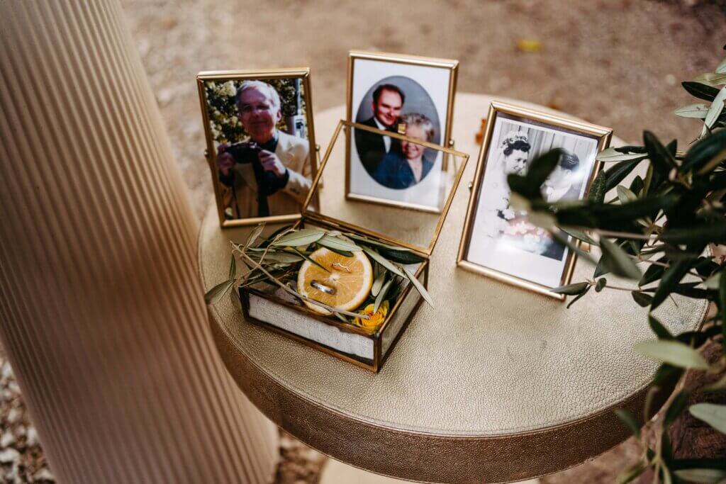 Auf einem runden Tisch stehen drei gerahmte Fotos von Menschen, ein kleiner Glaskasten und ein mit getrockneten Orangenscheiben und Blättern geschmücktes Buch. Der Hintergrund ist ein Außenbereich mit einer Säule und etwas Grün in der Nähe.