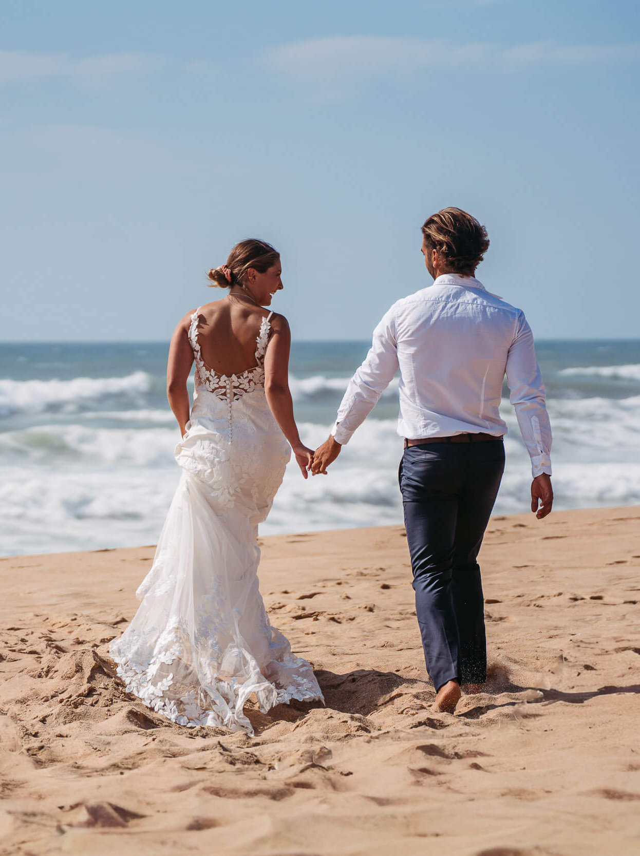 Eine Braut in einem weißen Hochzeitskleid und ein Bräutigam in einem weißen Hemd und einer dunklen Hose gehen Hand in Hand an einem Sandstrand entlang in Richtung Meer unter einem klaren Himmel.